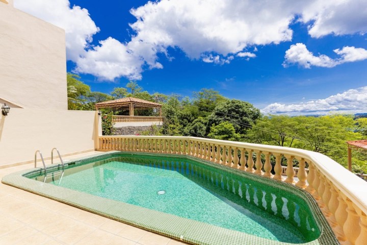 Sunny patio with a small pool surrounded by a balustrade, overlooking lush green trees and a clear blue sky.