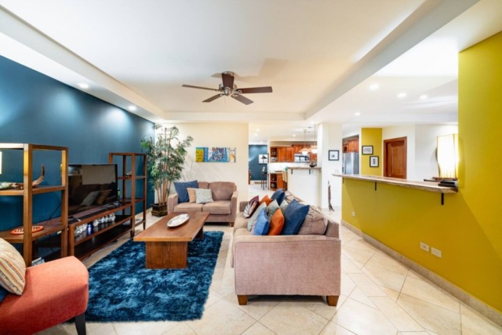 Modern living room with beige sofas, blue accent wall, ceiling fan, and open kitchen area.