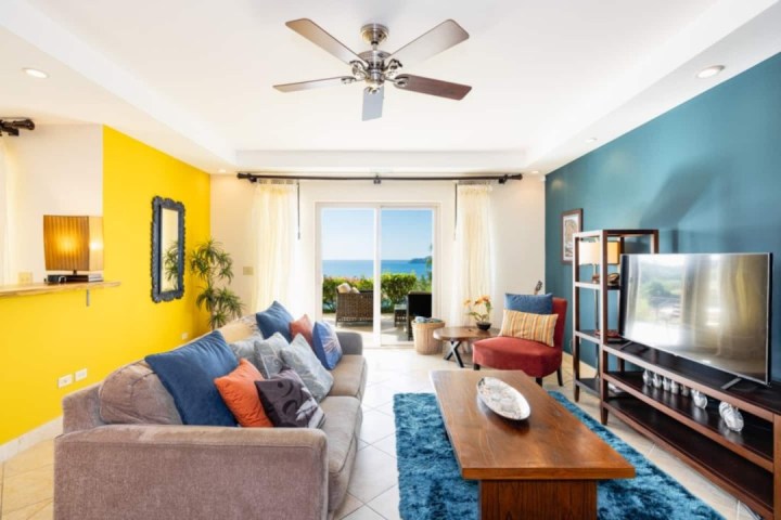 Bright living room with yellow and teal walls, sofa, chair, TV, ceiling fan, and ocean view through sliding doors.
