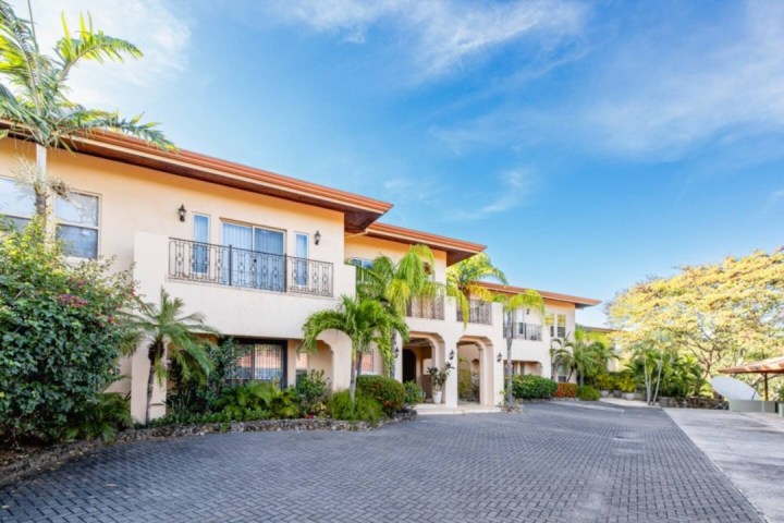 A two-story building with balconies, surrounded by palm trees and a paved driveway under a clear blue sky.