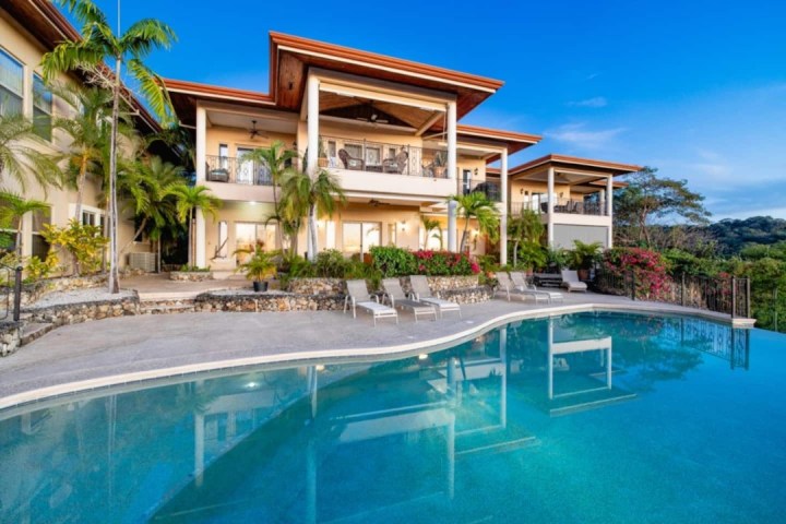 Luxury villa with large pool, palm trees, and balcony under a clear blue sky.