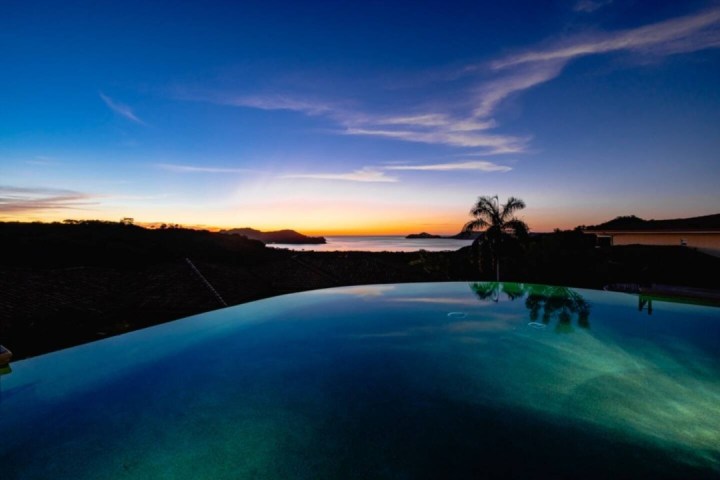 Infinity pool at sunset overlooking ocean with palm trees and hilly landscape.