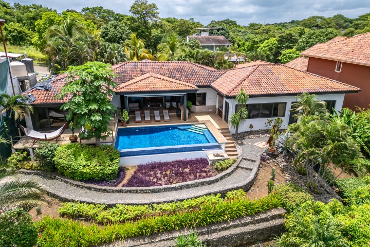 Aerial view of a house with a red-tiled roof, pool, and lush garden amidst trees.