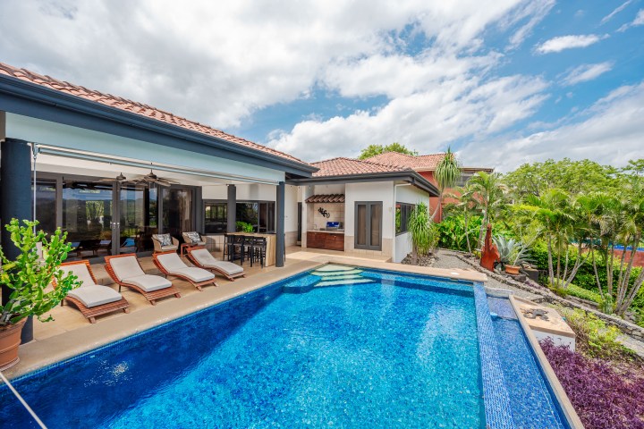 Poolside view of a modern house with lounge chairs, palm trees, and clear blue sky.