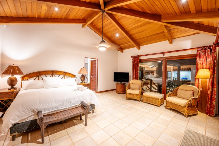 Spacious bedroom with wooden ceiling, bed, chairs, and a TV on tiled floor, featuring warm lighting.