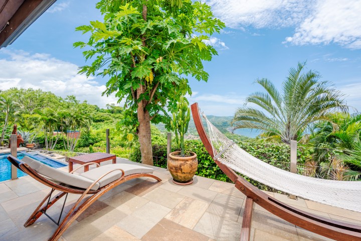 Tropical patio with hammock and loungers, overlooking a lush garden and ocean view under a blue sky.