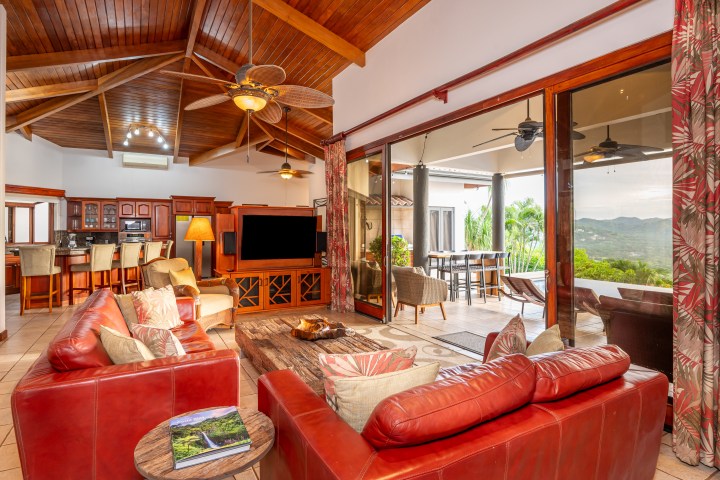 Spacious living room with red sofas, wooden ceiling, open kitchen, and view of patio with mountains.