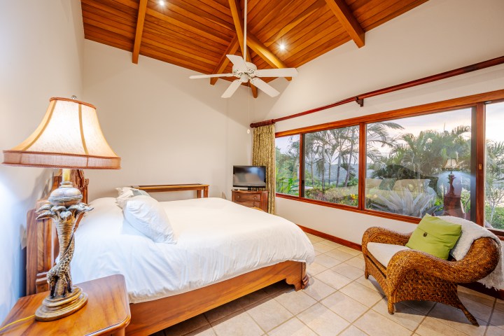 Cozy bedroom with wooden ceiling, large window, bed, wicker chair, lamp, and tropical garden view.