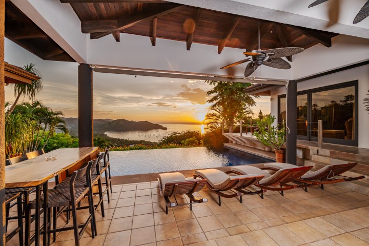 Outdoor patio with infinity pool, lounge chairs, sunset view over ocean and islands.