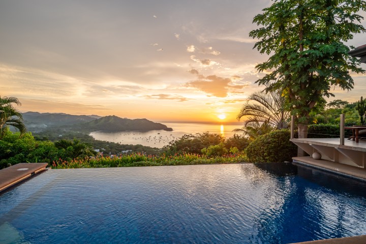Infinity pool overlooking ocean bay at sunset with lush greenery.