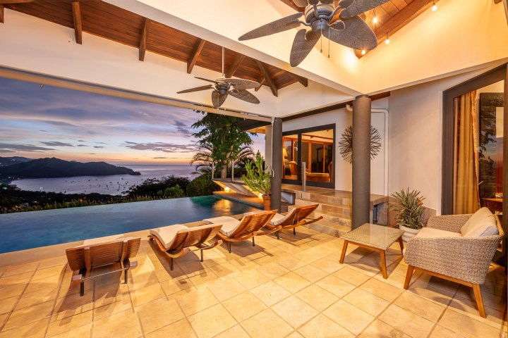 Luxurious patio with infinity pool overlooking ocean at sunset, featuring lounge chairs and ceiling fans.