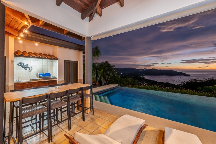 Outdoor patio with bar, pool, ocean view, and sunset sky.