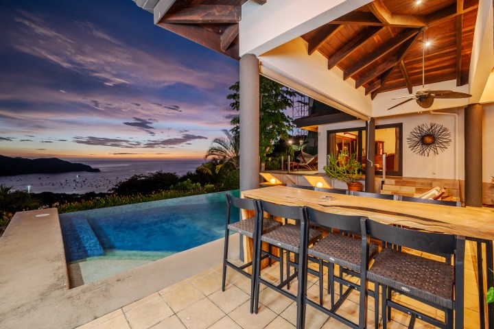 Covered patio with wooden table and chairs, pool, and ocean view at sunset.