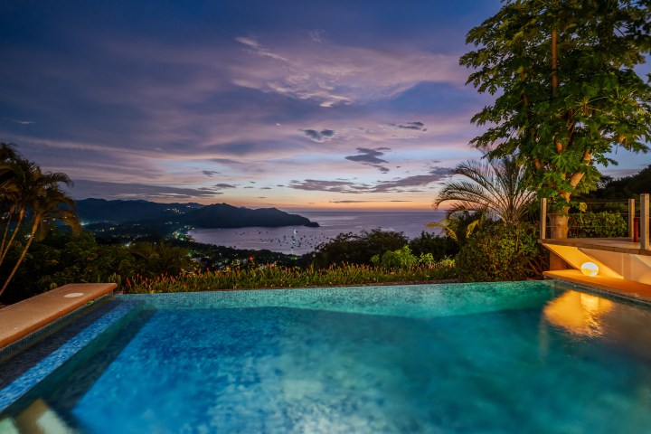 Infinity pool with ocean view at dusk, surrounded by trees and hills.