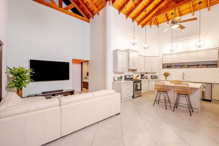 Open living room and kitchen with modern decor, white sofa, TV, island with stools, and wooden ceiling.