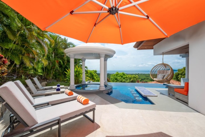 Poolside lounge with orange umbrella, deck chairs, swing chair, ocean views, and greenery.