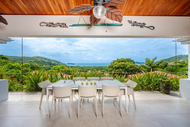 Outdoor dining area with ocean view, white chairs, table, and ceiling fan, surrounded by lush greenery.
