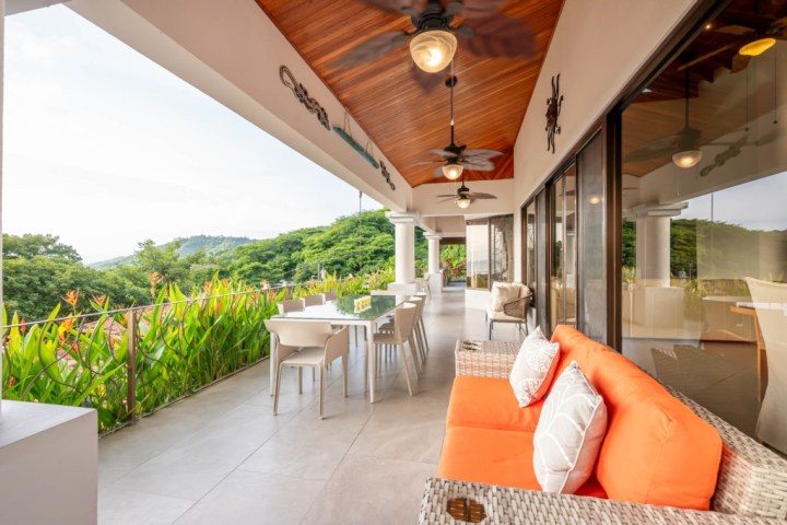Modern patio with orange sofa, dining table, ceiling fans, and lush greenery view.
