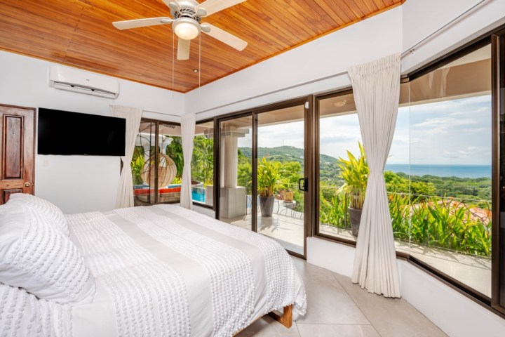 Bright bedroom with ceiling fan, large windows showing ocean view, and a wall-mounted TV.