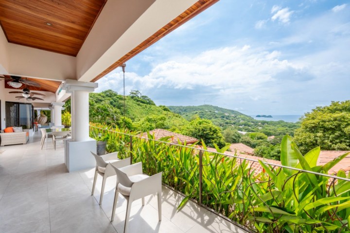 Spacious balcony with chairs, overlooking lush green hills and ocean view under a cloudy sky.