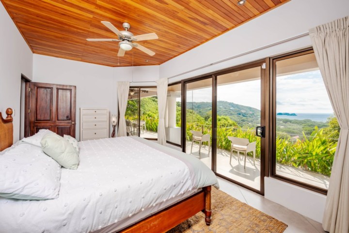 Cozy bedroom with wooden ceiling and large windows revealing a lush mountain and ocean view.