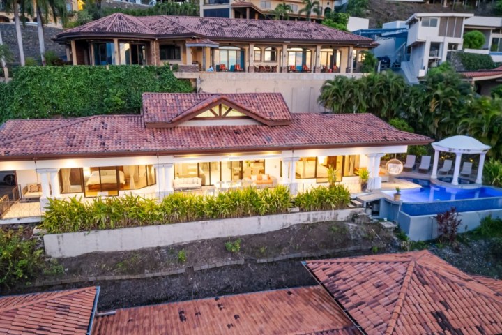 Aerial view of a luxurious hillside villa with red-tiled roof, bright lights, and an infinity pool.