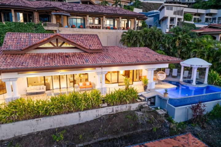 Luxurious villa with infinity pool and gazebo, surrounded by lush greenery and other homes on a hillside.