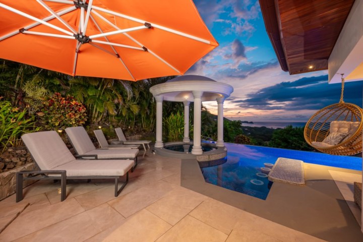 Patio with loungers, pool, umbrella, swing chair, and ocean view at sunset.