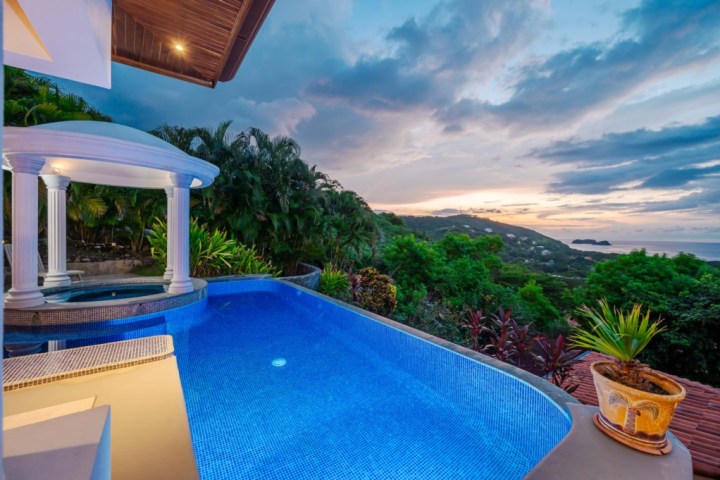 Infinity pool with ocean view at sunset, surrounded by trees and columns.
