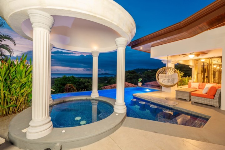 Luxurious outdoor pool area with columns, hot tub, and cozy seating at sunset.