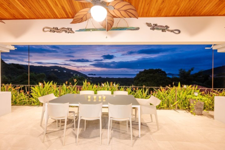 Outdoor dining area with white chairs, table, and sunset view of hills and sea.