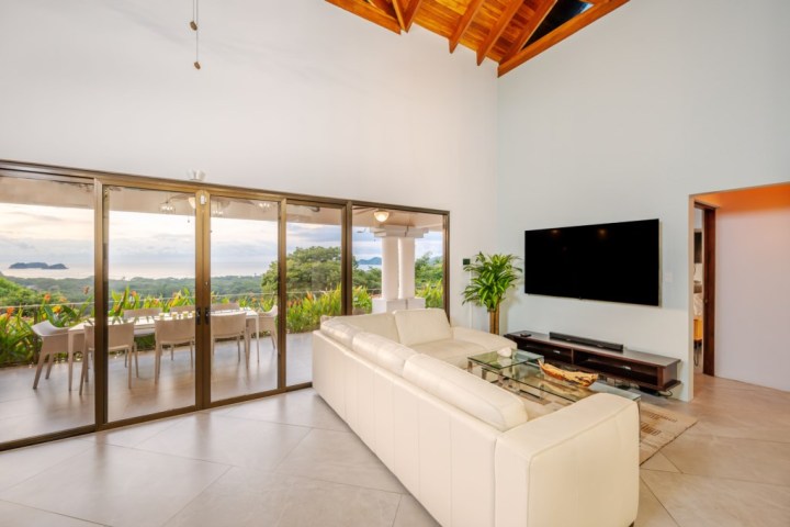 Modern living room with white sofa, glass coffee table, large TV, and ocean-view terrace.