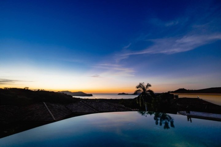 Infinity pool reflecting a vibrant sunset over hills and ocean, with palm trees silhouetted in the distance.