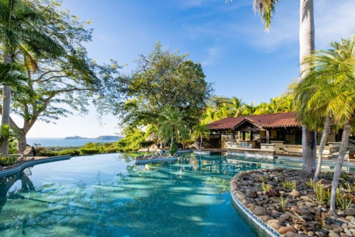 Tropical pool area with palm trees, a small pavilion, and ocean view under a clear blue sky.