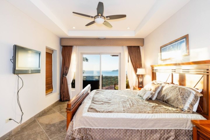 Bedroom with bed, TV, ceiling fan, and sea view through glass doors.