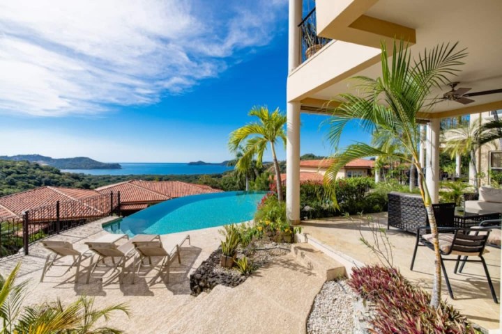 Luxurious patio with infinity pool, ocean view, palm trees, and lounge chairs under blue sky.