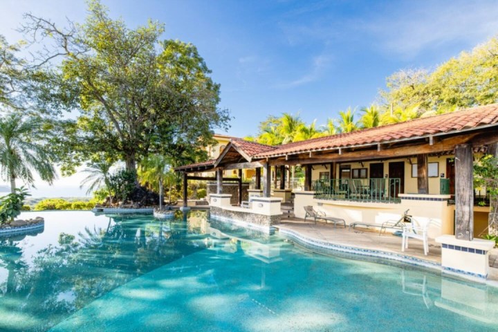 Tropical pool and patio with trees and lounge chairs, under a clear blue sky.