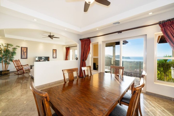 Dining room with wooden table, chairs, and ocean view through large windows, ceiling fan above.