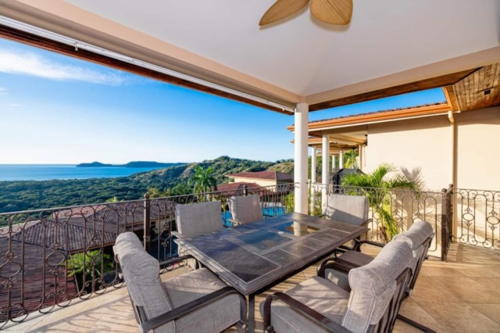 Outdoor patio with table and chairs overlooking ocean and hills.