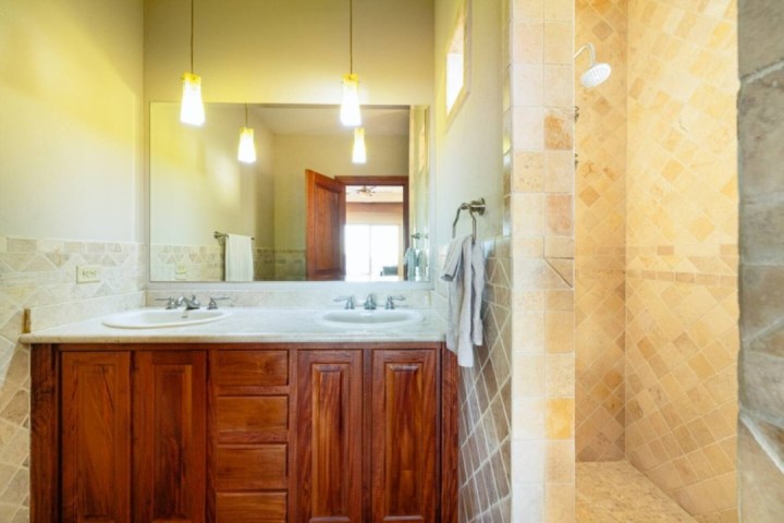Bathroom with double sink, wooden vanity, large mirror, hanging lights, and tiled shower area.