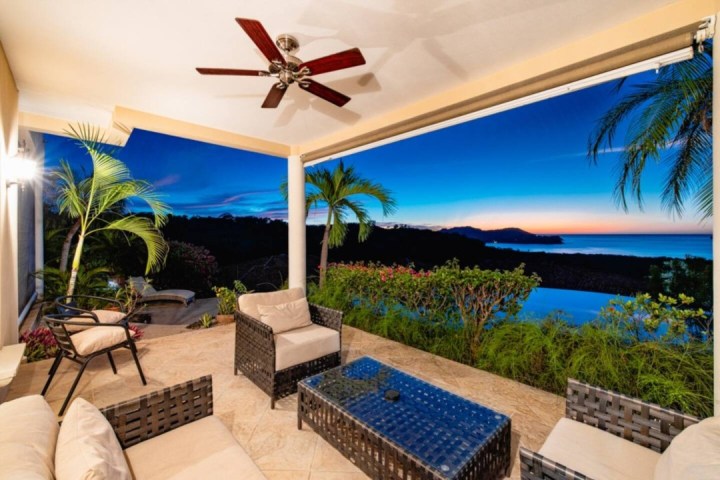 Outdoor patio with seating, palm trees, and ocean view at sunset.