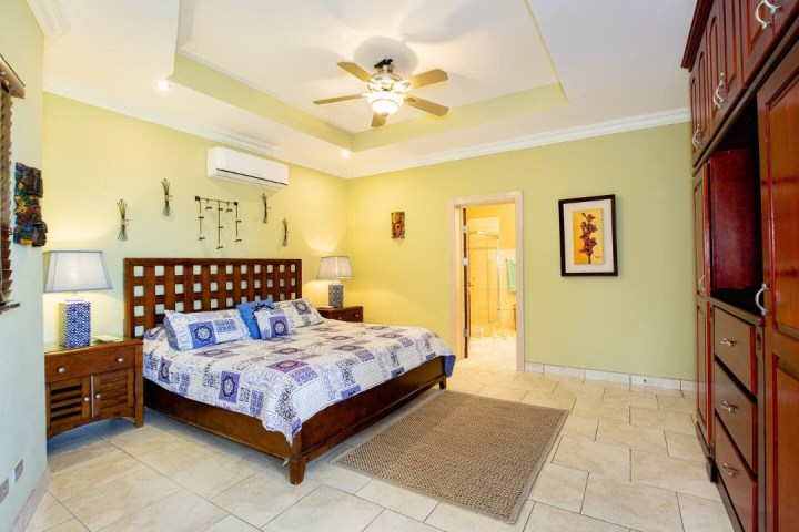 Bedroom with a large bed, ceiling fan, and adjacent bathroom. Walls are light yellow with decorative art.