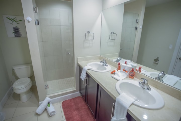 Bathroom with double sink, glass shower, toilet, towels, and toiletries.