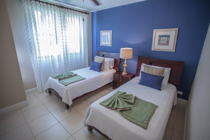 Room with two twin beds, blue wall, bedside lamp, and framed artwork.