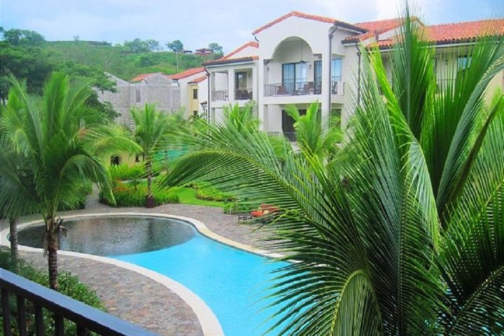 Tropical resort with pool, palm trees, and buildings with red roofs.