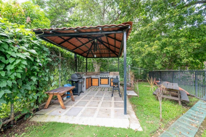 Outdoor kitchen with grill under a covered gazebo, surrounded by greenery and picnic table nearby.