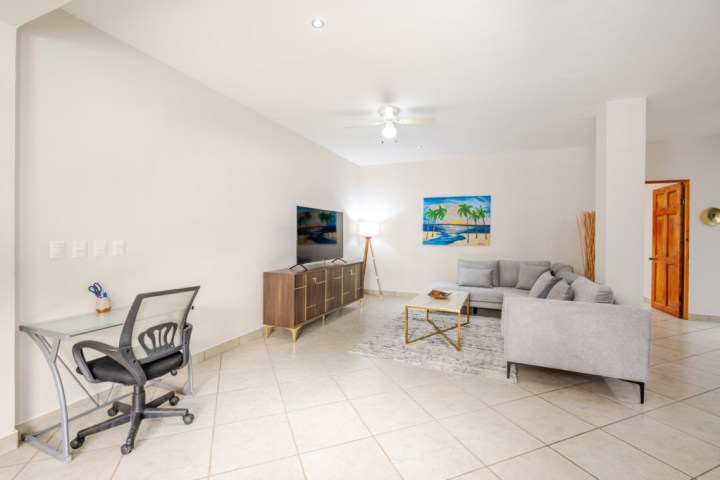 Modern living room with gray sofa, glass desk, TV, and palm tree painting on the wall.