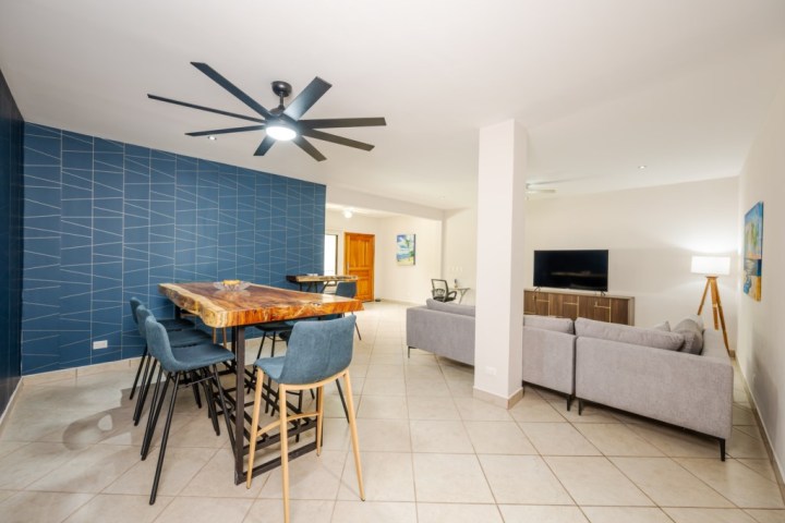 Modern living room with blue accent wall, wooden dining table, gray sofa, and TV.