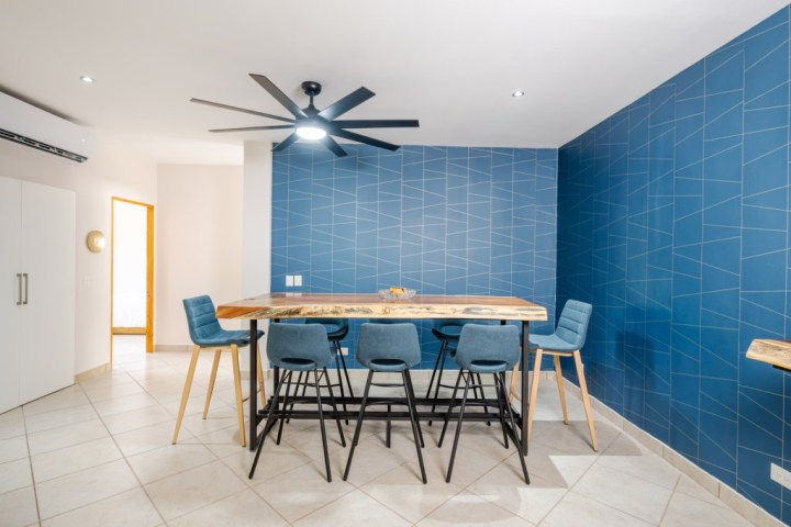 Modern dining area with wooden table, blue chairs, blue geometric wall, and ceiling fan.