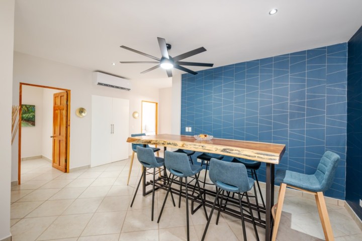 Modern dining room with blue accent wall, wooden table, and blue chairs under a ceiling fan.